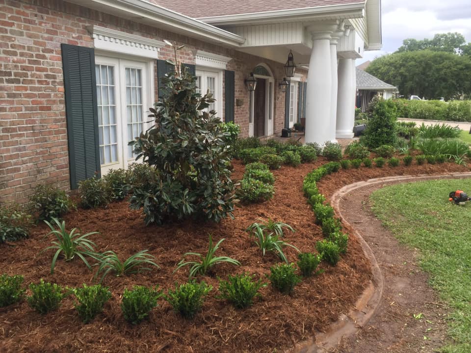 Freshly mulched garden beds with neatly planted greenery along a curved brick border in front of a classic brick home, maintained by Eric's Tidy Turf in Gretna, LA