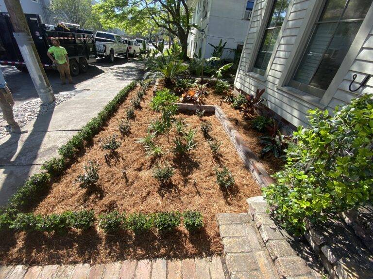 Beautifully landscaped front garden bed with vibrant purple foliage and green shrubs, framed by a brick border in front of a home, maintained by Eric's Tidy Turf.