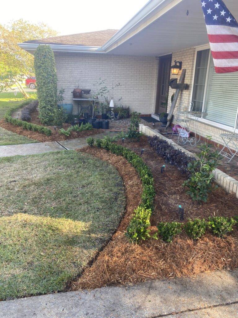 Beautifully landscaped front garden bed with vibrant purple foliage and green shrubs, framed by a brick border in front of a home, maintained by Eric's Tidy Turf.