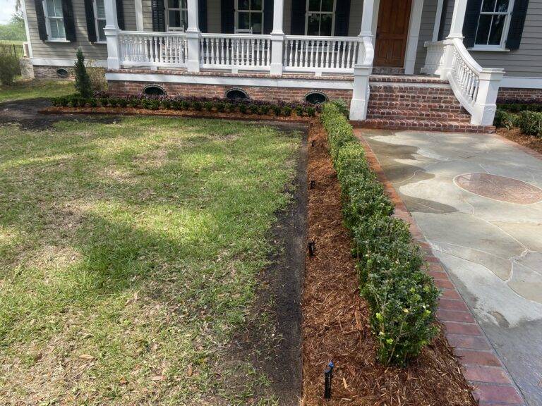 Beautifully landscaped front garden bed with vibrant purple foliage and green shrubs, framed by a brick border in front of a home, maintained by Eric's Tidy Turf.