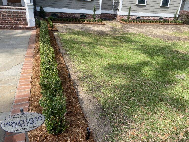 Beautifully landscaped front garden bed with vibrant purple foliage and green shrubs, framed by a brick border in front of a home, maintained by Eric's Tidy Turf.