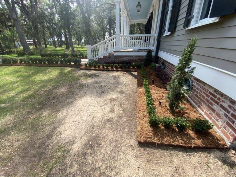 Beautifully landscaped front garden bed with vibrant purple foliage and green shrubs, framed by a brick border in front of a home, maintained by Eric's Tidy Turf.