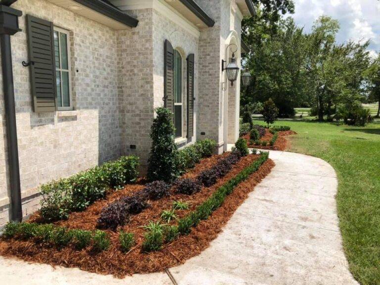 Beautifully landscaped front garden bed with vibrant purple foliage and green shrubs, framed by a brick border in front of a home, maintained by Eric's Tidy Turf.