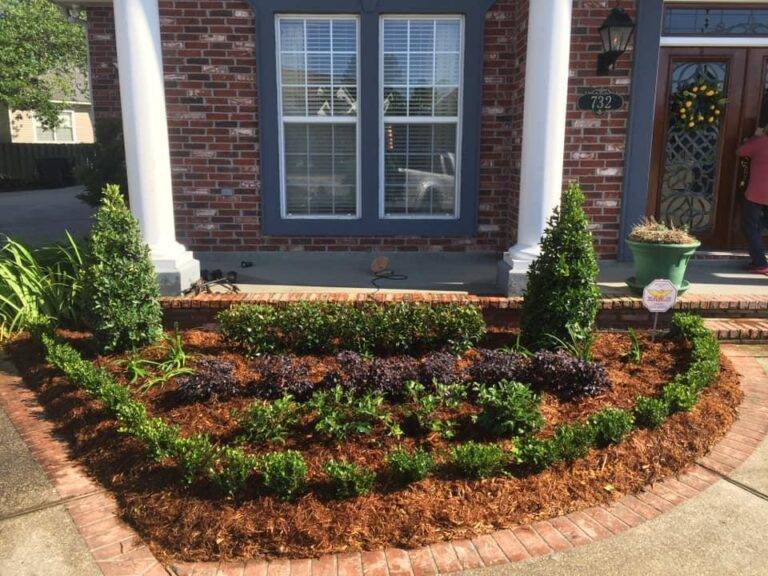 Beautifully landscaped front garden bed with vibrant purple foliage and green shrubs, framed by a brick border in front of a home, maintained by Eric's Tidy Turf.