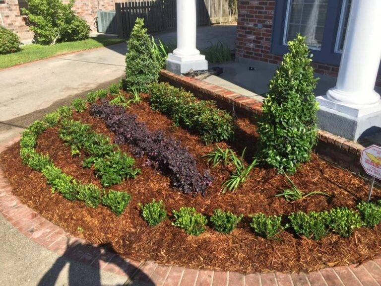 Beautifully landscaped front garden bed with vibrant purple foliage and green shrubs, framed by a brick border in front of a home, maintained by Eric's Tidy Turf.