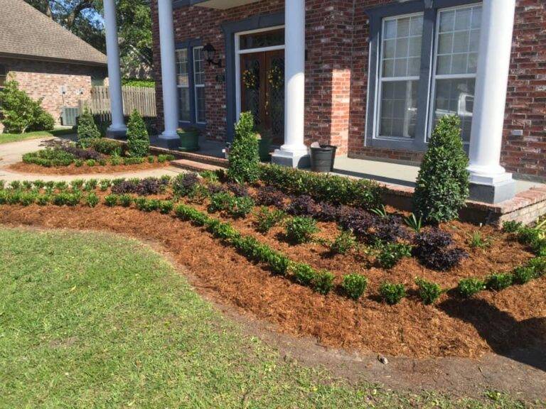 Beautifully landscaped front garden bed with vibrant purple foliage and green shrubs, framed by a brick border in front of a home, maintained by Eric's Tidy Turf.